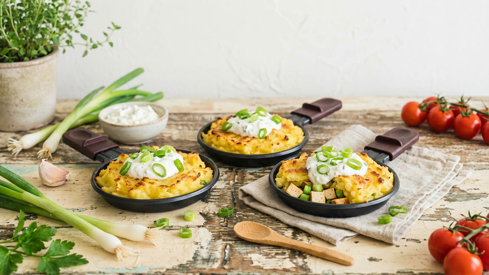 Drei kleine Pfännchen mit gebackenem Kartoffelbrei sind auf einem Holztisch angeordnet. Jedes Pfännchen ist mit Sauerrahm und frischen, gehackten Frühlingszwiebeln garniert. Im Hintergrund sind frische Kräuter, eine Schale mit Quark und einige Cherry-Tomaten zu sehen.