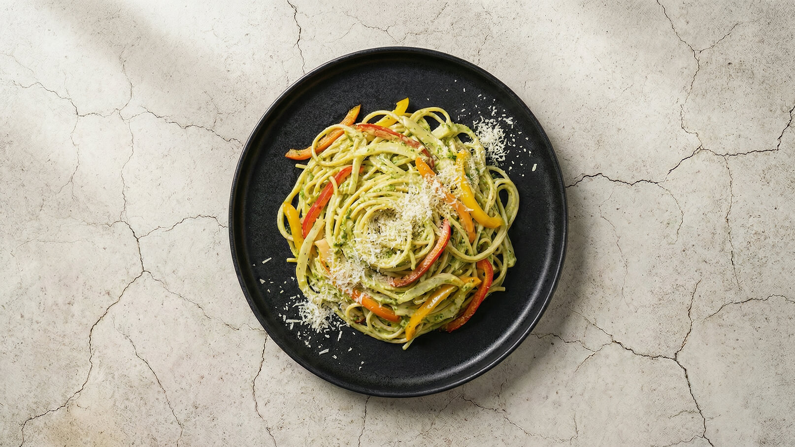 Ein schwarzer Teller mit einer Portion Spaghetti, die mit einer grünen Sauce und buntem Gemüse, wie roten und gelben Paprika, serviert sind. Darüber streut ein feiner Käse. Der Hintergrund hat eine helle, strukturierte Oberfläche.