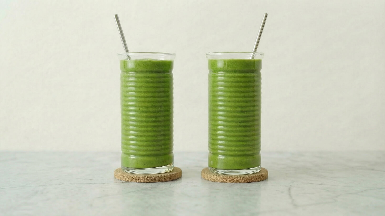 Zwei hohe Gläser mit grünem Smoothie stehen auf runden Untersetzern. Jedes Glas hat einen Strohhalm und ist leicht gerippt. Der Hintergrund ist hell und strukturiert, der Tisch fühlt sich glatt an.