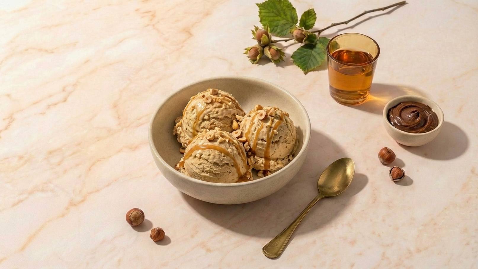 Eine Schüssel mit drei Kugeln Nusseis, garniert mit Nüssen und Karamellsoße. Daneben ein kleiner Teller mit Schokoladensauce, eine Gabel und ein Glas mit einem amberfarbenen Getränk. Einige Haselnüsse liegen ebenfalls auf dem Tisch.