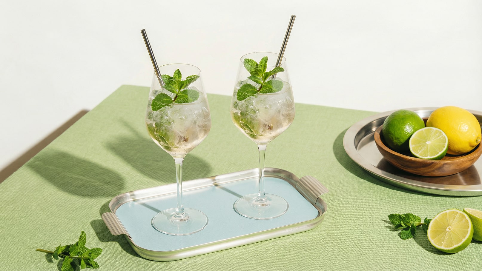 Zwei Gläser mit erfrischendem Getränk, gefüllt mit Eis und frischer Minze, stehen auf einem blauen Tablett. Im Hintergrund liegen eine Schale mit Zitronen und Limetten sowie frische Minzblätter auf einem grünen Tisch.
