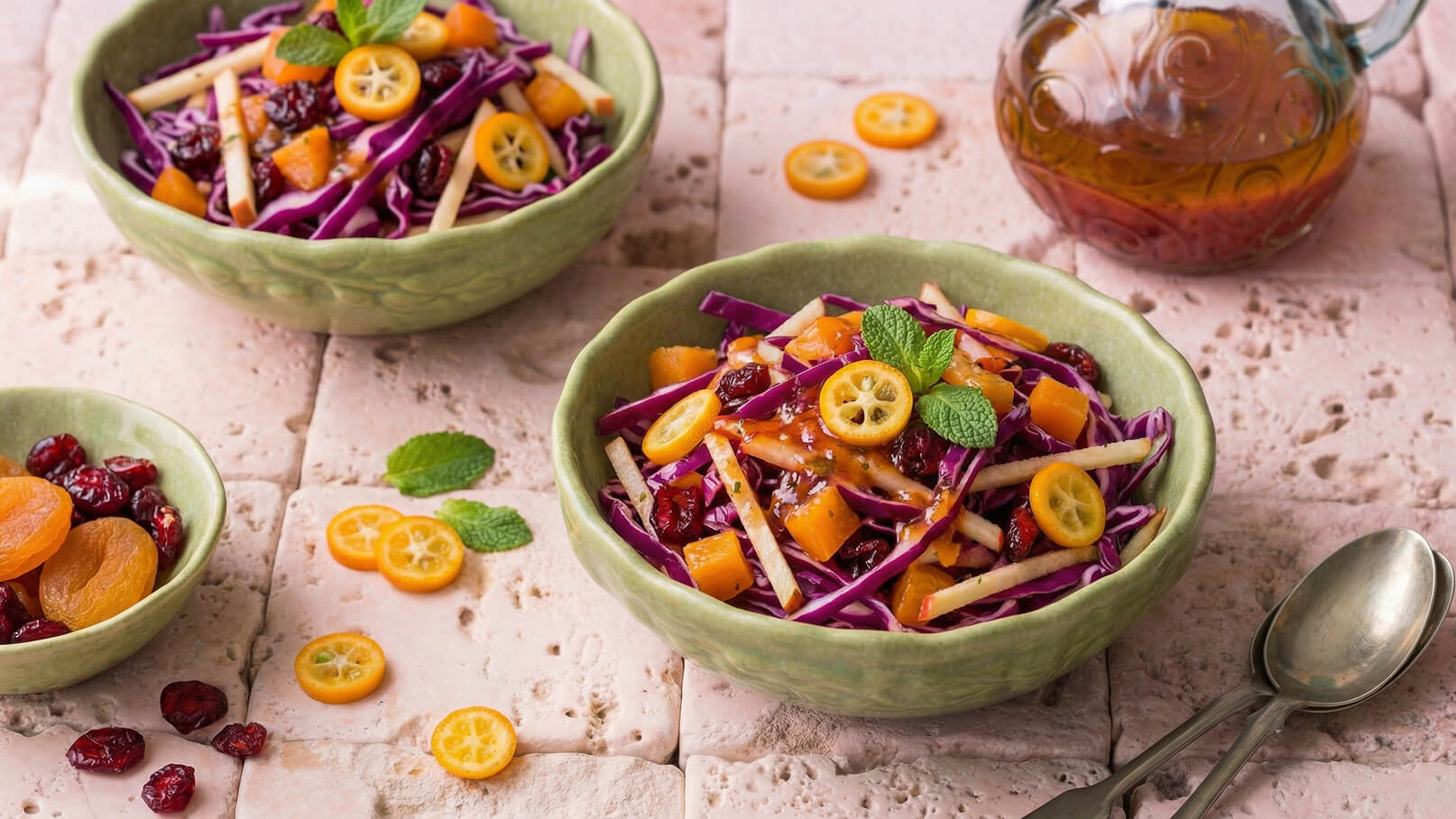 Zwei grüne Schalen mit buntem Salat aus rotem Kohl, Äpfeln, getrockneten Früchten und Kumquats. Frische Minzblätter sind als Garnitur auf dem Salat platziert. Im Hintergrund steht eine Glasflasche mit Dressing. Der Tisch hat eine rustikale Steinoberfläche.