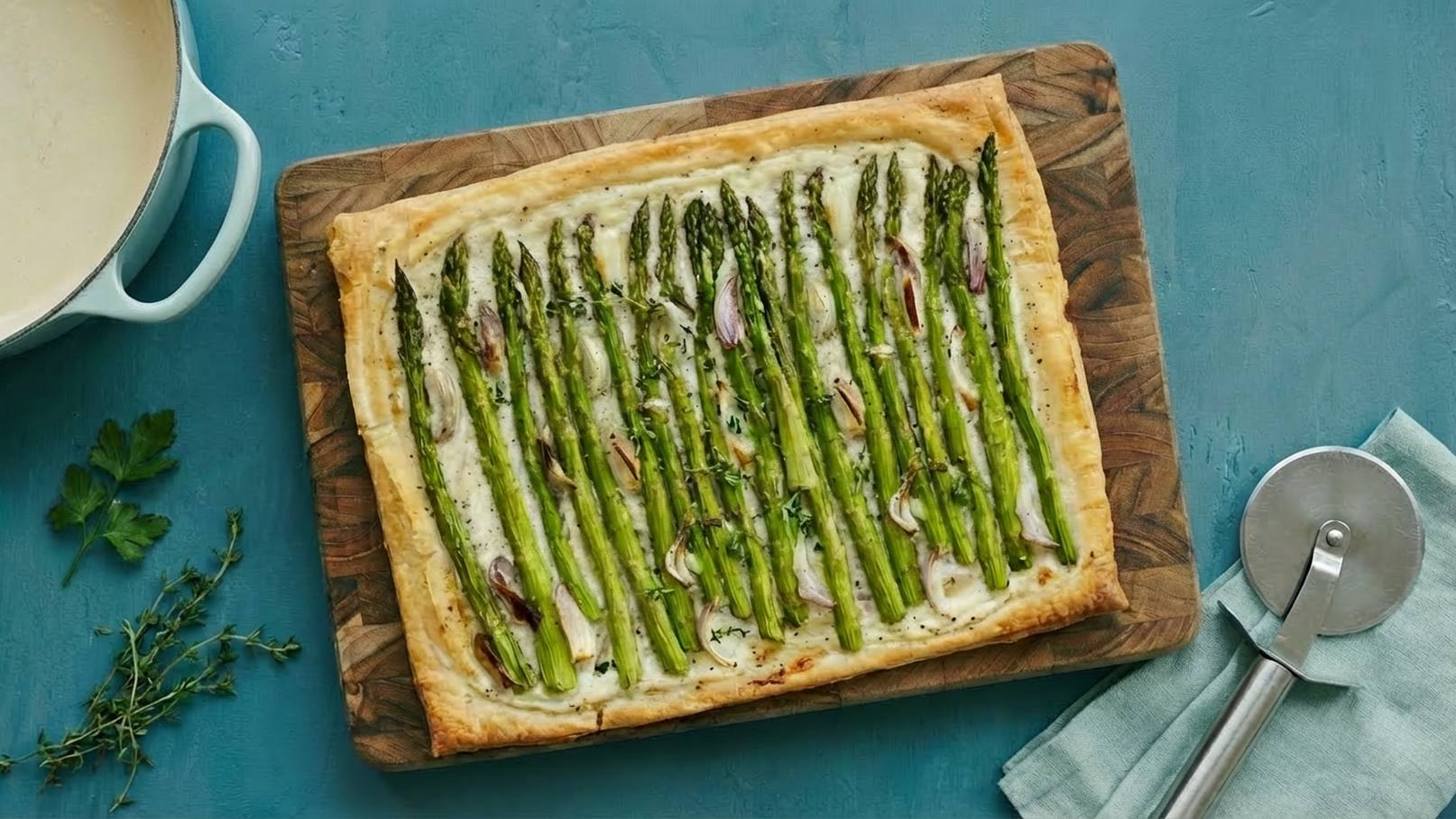 Ein frisch gebackener Blätterteig mit grünen Spargelstangen, garniert mit Zwiebeln und Kräutern. Der Kuchen liegt auf einem Holzbrett. Im Hintergrund ist eine helle Schüssel mit einer cremigen Sauce sichtbar.