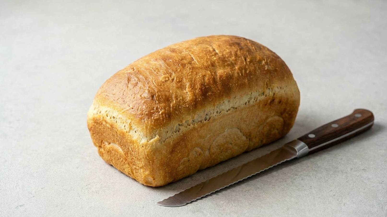 Ein frisch gebackenes, goldbraunes Brotlaib liegt auf einer hellen Oberfläche, daneben liegt ein gezacktes Brotmesser mit einem Holzgriff.