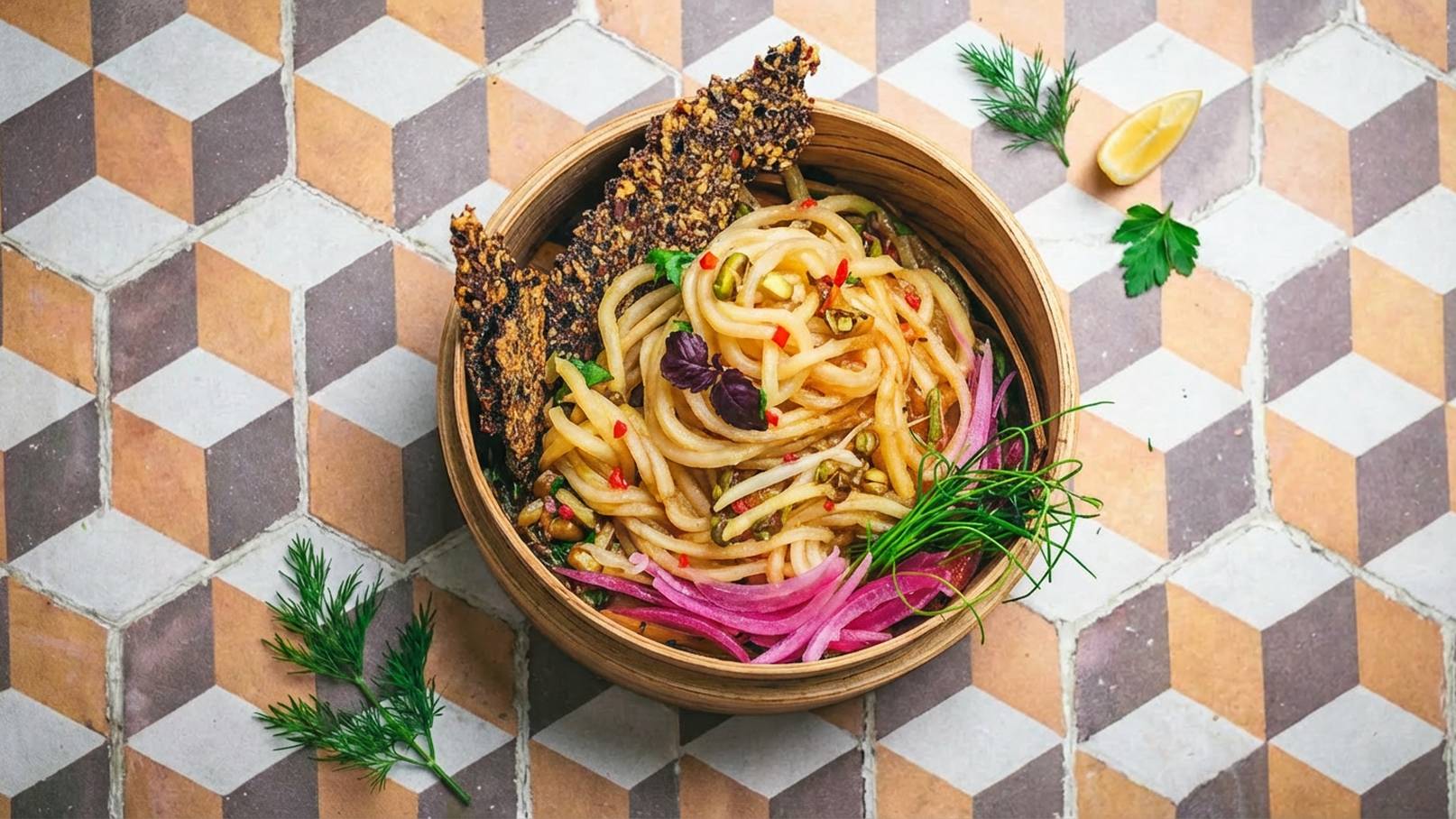 Eine Holzschüssel mit bunten Nudeln, garniert mit roten Zwiebeln, frischen Kräutern und einer knusprigen Beilage aus Sesam. Die Nudeln sind mit verschiedenen Gewürzen und möglicherweise einem Hauch Zitrone verfeinert. Der Hintergrund besteht aus geometrischen Fliesen.