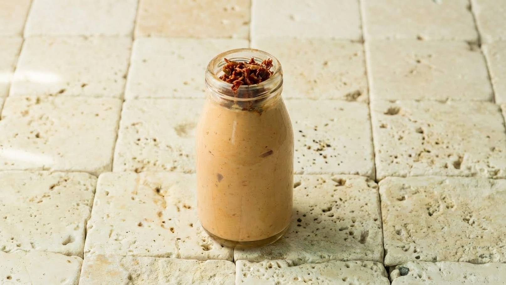 Ein Glas mit einer cremigen, beige-farbenen Paste ist auf einem strukturierten, hellen Untergrund platziert. Die Paste ist mit einer Schicht von knusprigen, braunen Stückchen oben dekoriert, die dekorativ wirken.