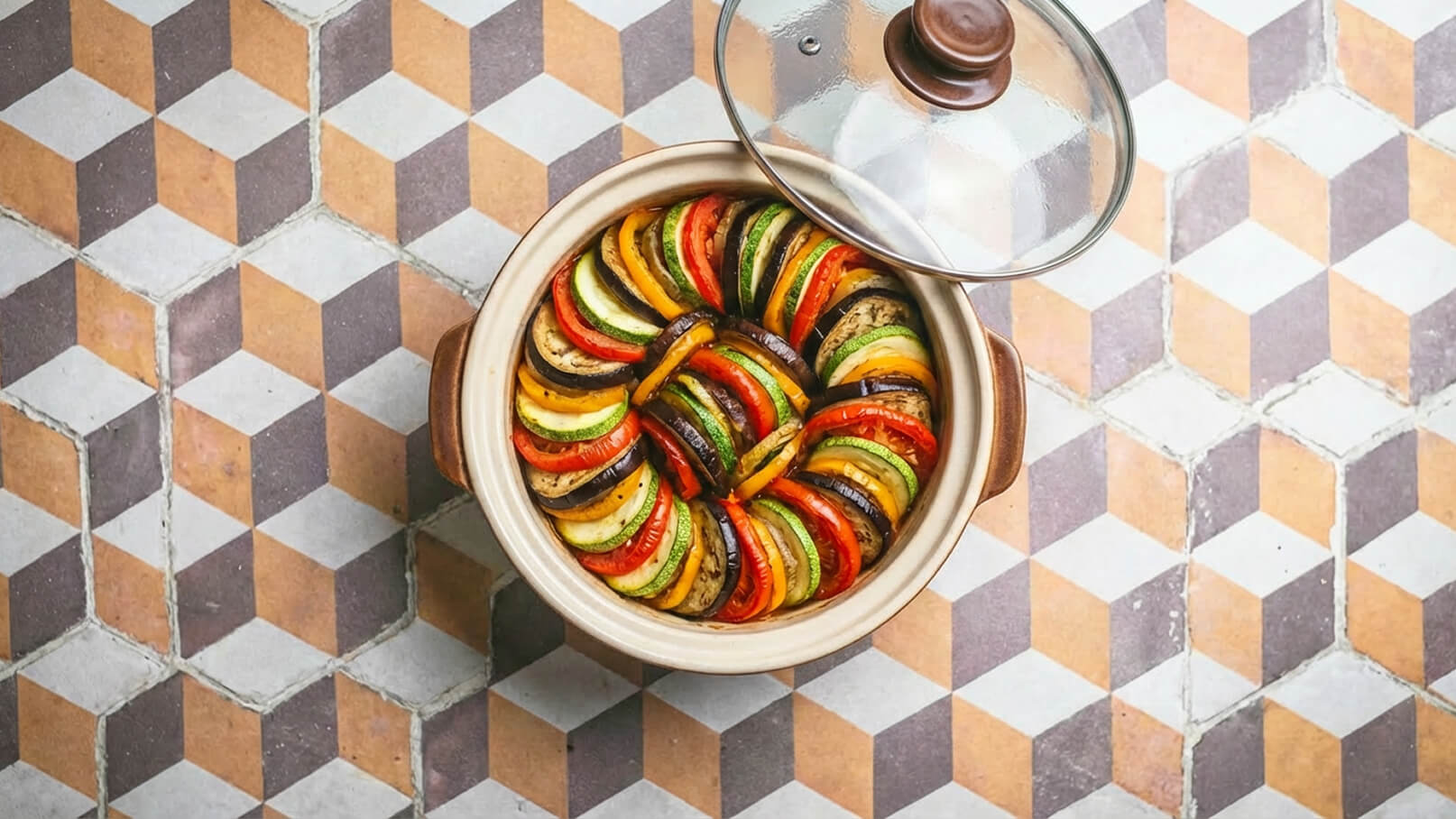 Ein runder Schmortopf mit kunstvoll geschichteten Gemüsesorten, darunter Zucchini, Auberginen, Paprika und Tomaten. Der Deckel ist leicht geöffnet, und der Topf steht auf einem geometrisch gemusterten Boden. Die Farben der Zutaten strahlen frisch und einladend.