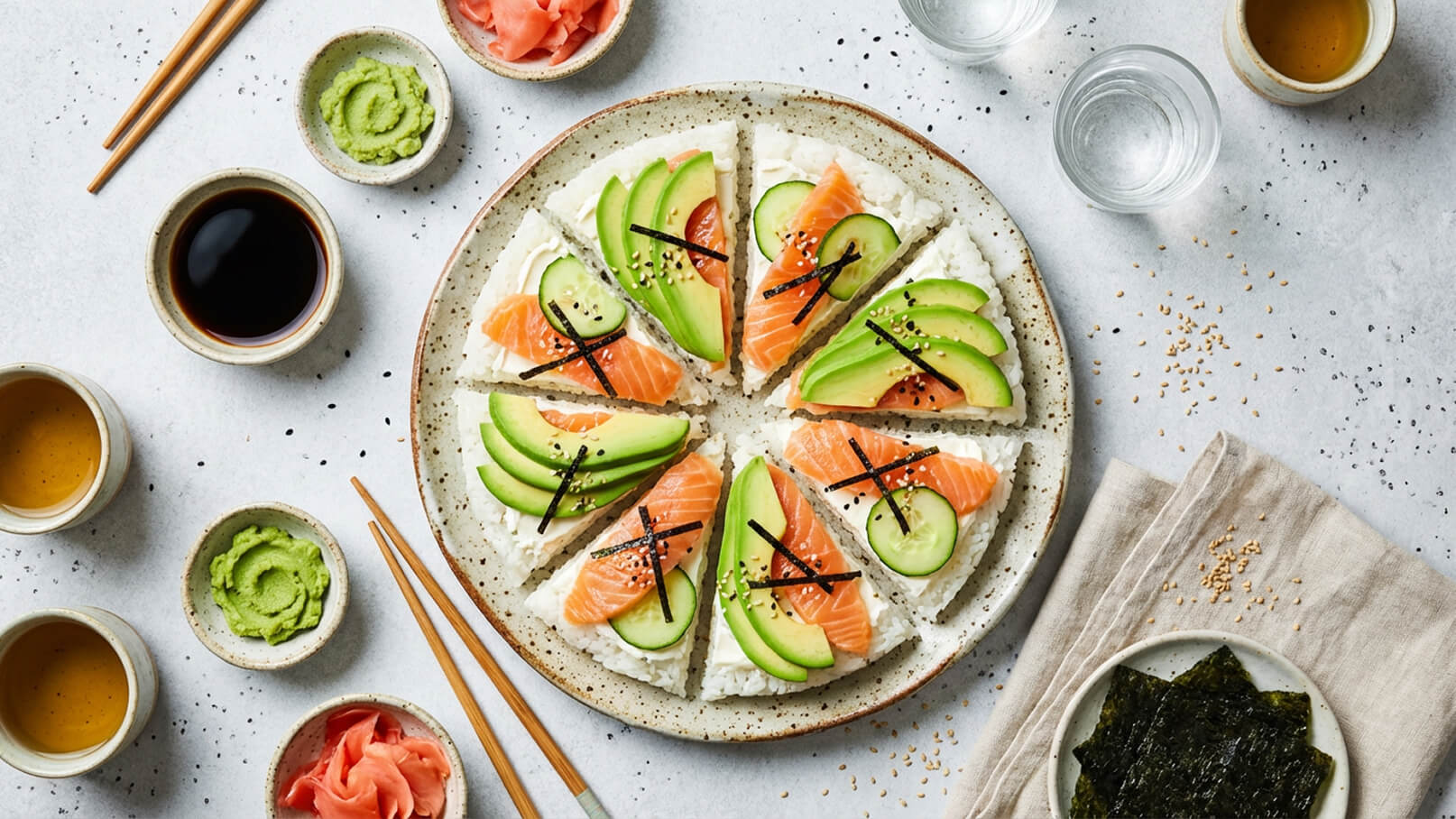 Ein runder Teller mit Sushi, aufgeteilt in Spalten. Jede Spalte ist belegt mit frischem Lachs, Gurkenscheiben und Avocado, garniert mit schwarzen Algenstreifen. Um den Teller sind kleine Schalen mit Sojasauce, Wasabi, Ingwer und Sesam verteilt, sowie Essstäbchen neben dem Teller.