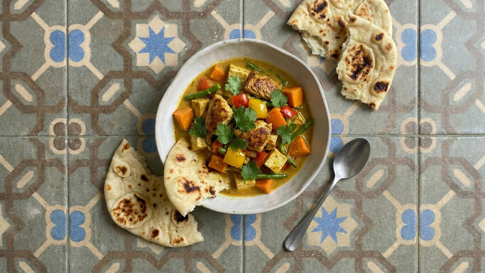 Eine Schüssel mit bunter Gemüsesuppe enthält Stücke von Tofu und verschiedenen Gemüsesorten wie Kürbis und Paprika, garniert mit frischem Koriander. Neben der Schüssel liegen einige Stücke Fladenbrot und ein Löffel auf einem bunten Musterboden.