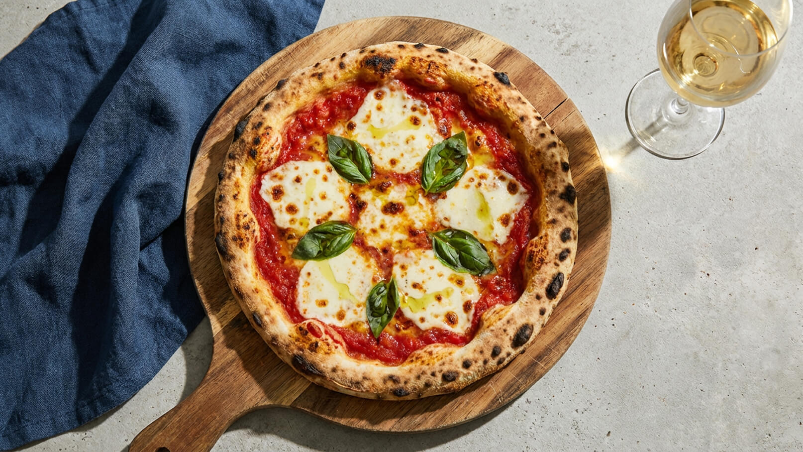 Eine frisch gebackene Pizza Margherita auf einem rustikalen Holzbrett. Die Pizza hat einen knusprigen Rand, ist mit Tomatensauce, Mozzarella und frischen Basilikumblättern belegt und wirkt appetitlich. Rechts im Bild steht ein Glas weißer Wein.