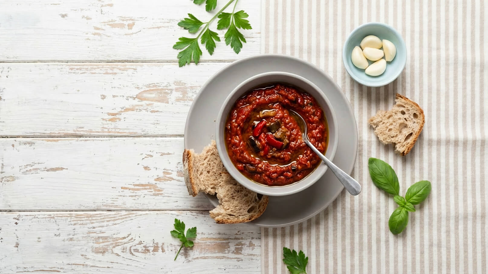 Eine Schüssel mit sämiger Tomatensuppe, garniert mit geschnittenem Gemüse, steht auf einem Teller. Neben der Schüssel liegen zwei Stücke Brot. Im Hintergrund sind ein paar Knoblauchzehen und frische Basilikumblätter zu sehen.