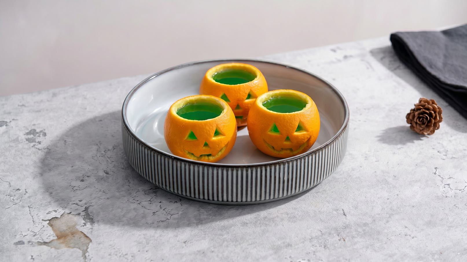 Auf einem weißen Teller befinden sich drei orangefarbene Halloween-Töpfe in Kürbisform. Die Töpfe haben eine grüne Innenseite und sind mit geschnittenen grünen Dreiecken verziert. Der Hintergrund ist schlicht und neutral.