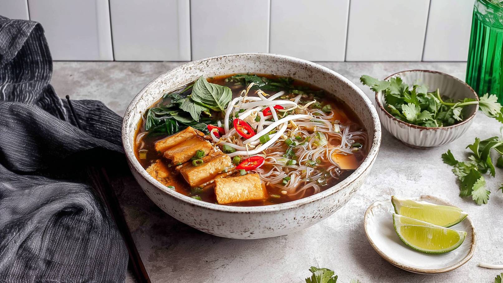 Eine Schüssel mit aromatischer Nudelsuppe, gefüllt mit Brühe, Reisnudeln, Tofu-Stücken, frischem Gemüse, Sprossen und geschnittenen roten Chilischoten. Neben der Suppe liegen frische Korianderblätter und Limettenscheiben auf einem kleinen Teller.
