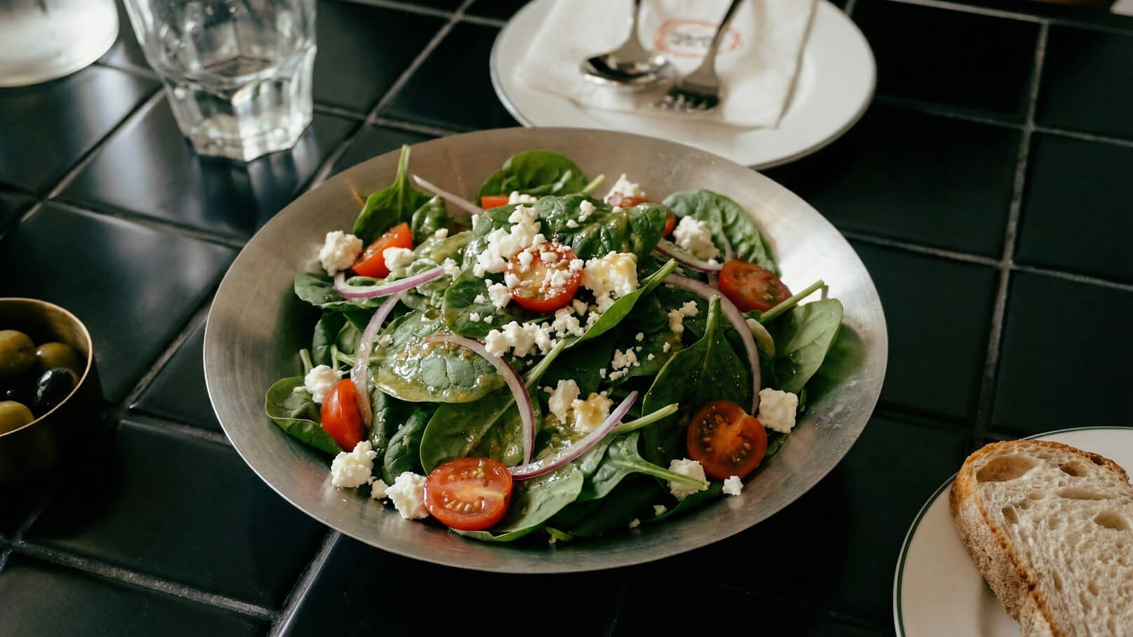 Ein silberner Teller enthält frischen Spinatsalat mit Kirschtomaten, roten Zwiebelringen und Feta-Käse. Neben dem Salat liegt ein Stück Brot. Der Hintergrund ist eine dunkle Tischoberfläche.