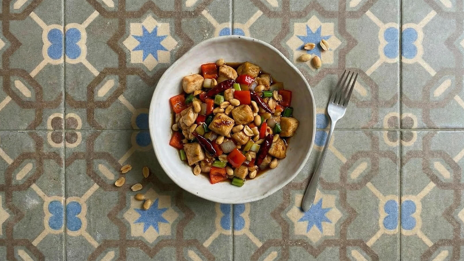 Eine weiße Schale mit buntem Gemüse, Hühnchenstücken und Erdnüssen steht auf einem gefliesten Tisch. Die Mischung enthält rote Paprika, Zwiebeln und grüne Frühlingszwiebeln. Neben der Schale liegt eine Gabel.
