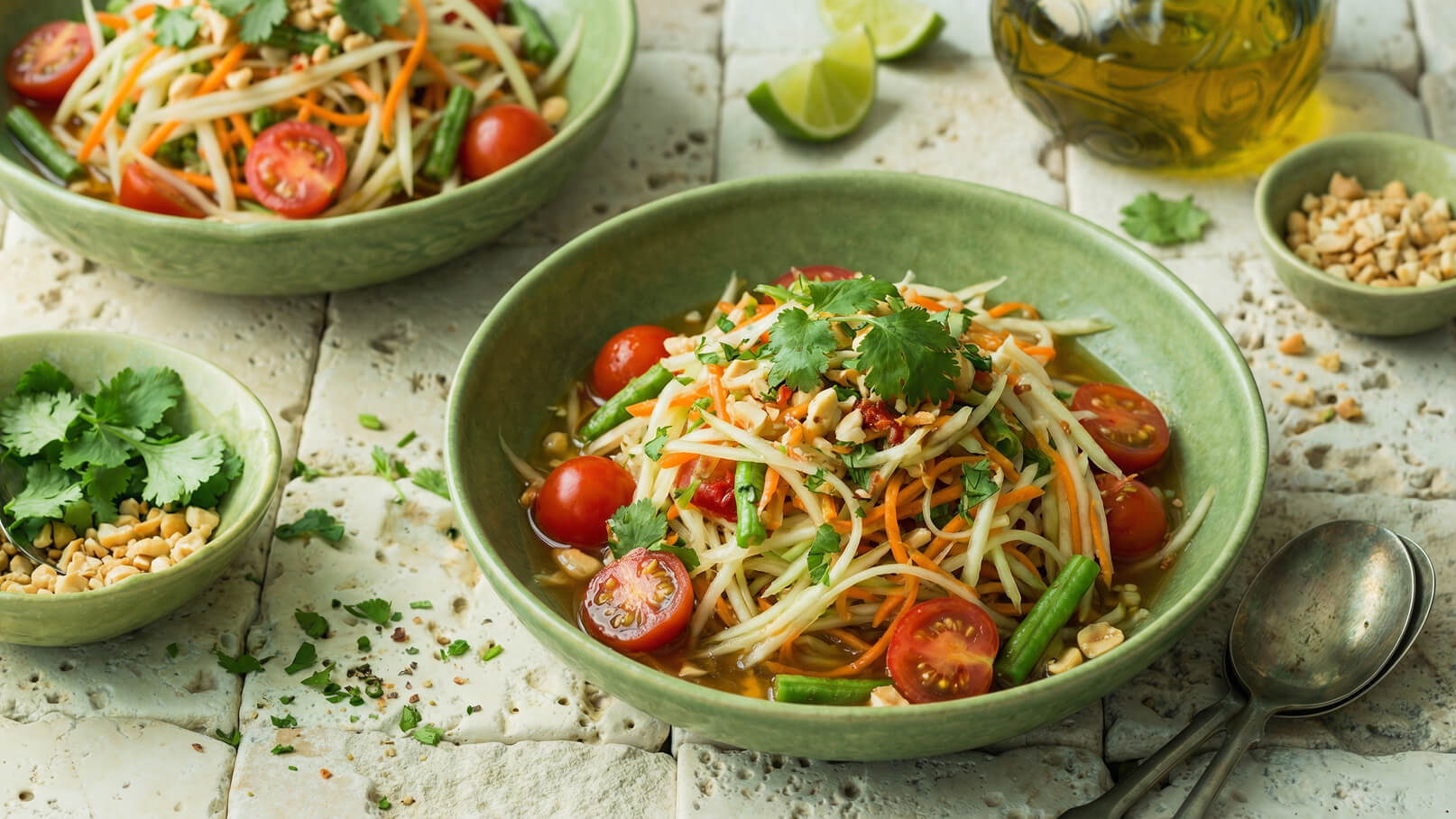 Zwei Schalen mit gefülltem Salat sind auf einem rustikalen Tisch angeordnet. Der Salat enthält grüne Bohnen, Karotten, Kirschtomaten und passt mit Koriander und Erdnüssen. Ein paar Limettenscheiben und Öl sind im Hintergrund sichtbar.