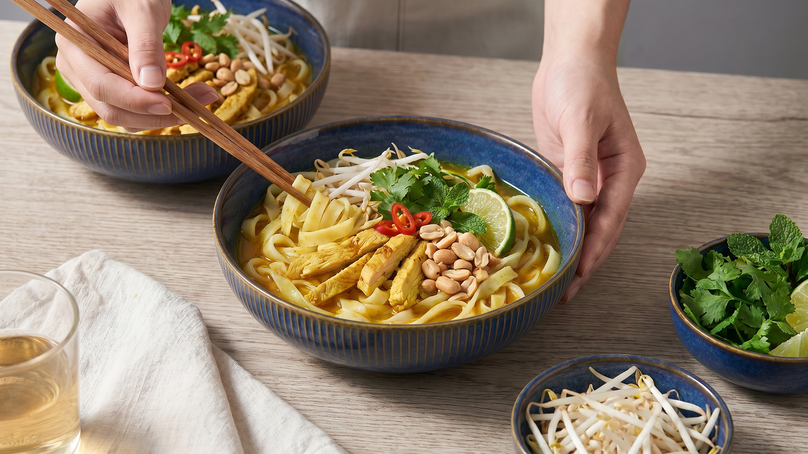 Zwei Schalen mit Nudeln, garniert mit Hähnchen, Erdnüssen, Limettenscheiben, frischem Koriander und roten Chiliflocken. Eine Hand hält Essstäbchen, um die Nudeln zu servieren. Neben den Schalen befinden sich Schalen mit Sprossen und frischen Kräutern sowie ein Glas Getränk.