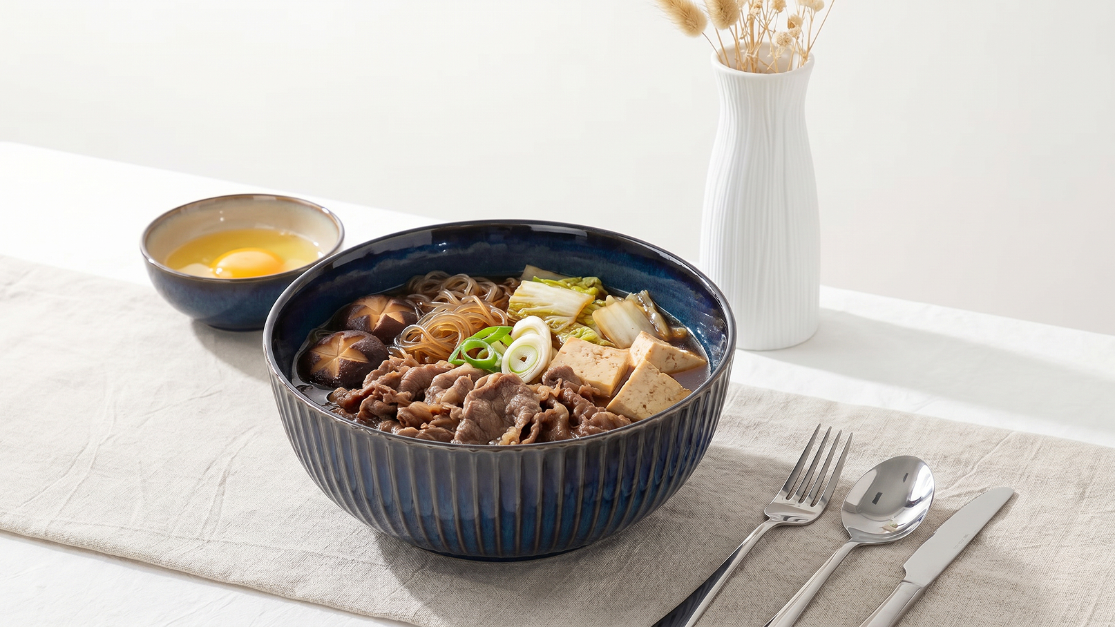 Eine Schüssel mit Ramen-Nudeln, belegt mit zartem Fleisch, Tofu, Frühlingszwiebeln und Gemüse. Daneben steht eine Schale mit einem rohen Ei und eine Vase mit Trockenblumen. Der Tisch ist einfach gedeckt mit Besteck und einer Tischdecke.