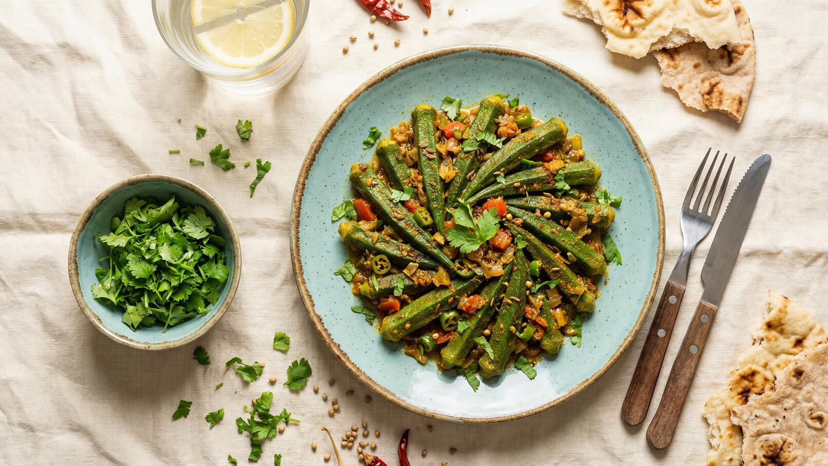 Ein Teller mit gebratenem Okra, garniert mit frischem Koriander und Tomatenwürfeln, steht auf einem Tisch. Daneben befindet sich eine Schüssel mit frischem Koriander, Besteck und etwas Brot. Im Hintergrund ist ein Glas Wasser mit einer Zitronenscheibe zu sehen.