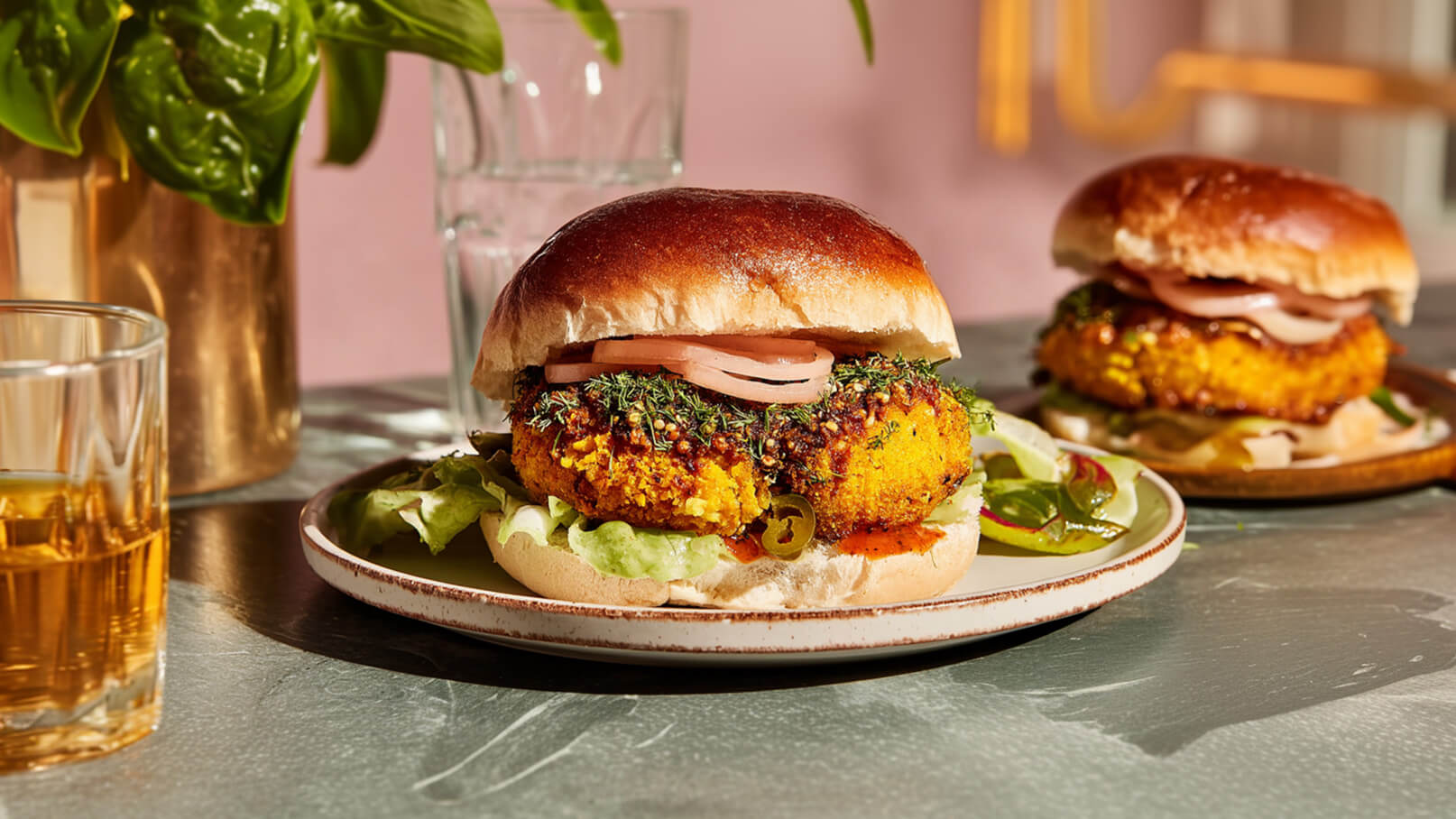 Auf dem Bild sind zwei Hamburger auf Tellern zu sehen. Jeder Burger besteht aus einem weichen Brötchen, einem knusprigen Patty, frischem Salat und einigen Zwiebelringen. Im Hintergrund befindet sich eine Pflanze und ein Glas Wasser.