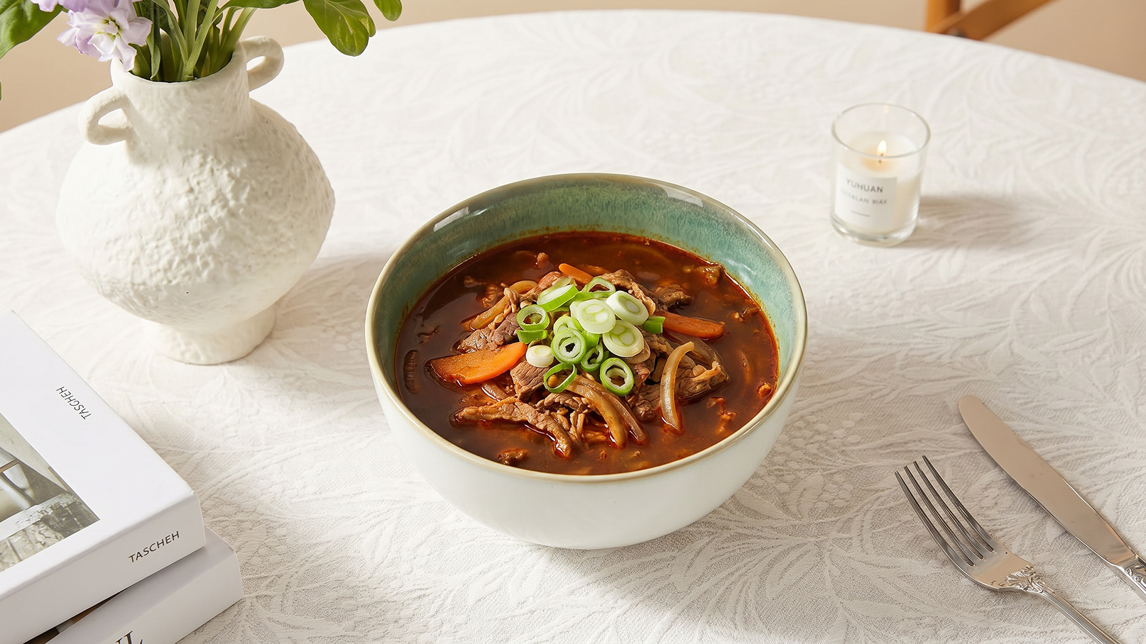 Eine Schüssel mit würziger Suppe steht auf einem gedeckten Tisch. Die Suppe enthält Stücke von Fleisch, Gemüse und ist mit geschnittenen Frühlingszwiebeln garniert. Im Hintergrund sind eine Blumenvase und eine Kerze sichtbar.