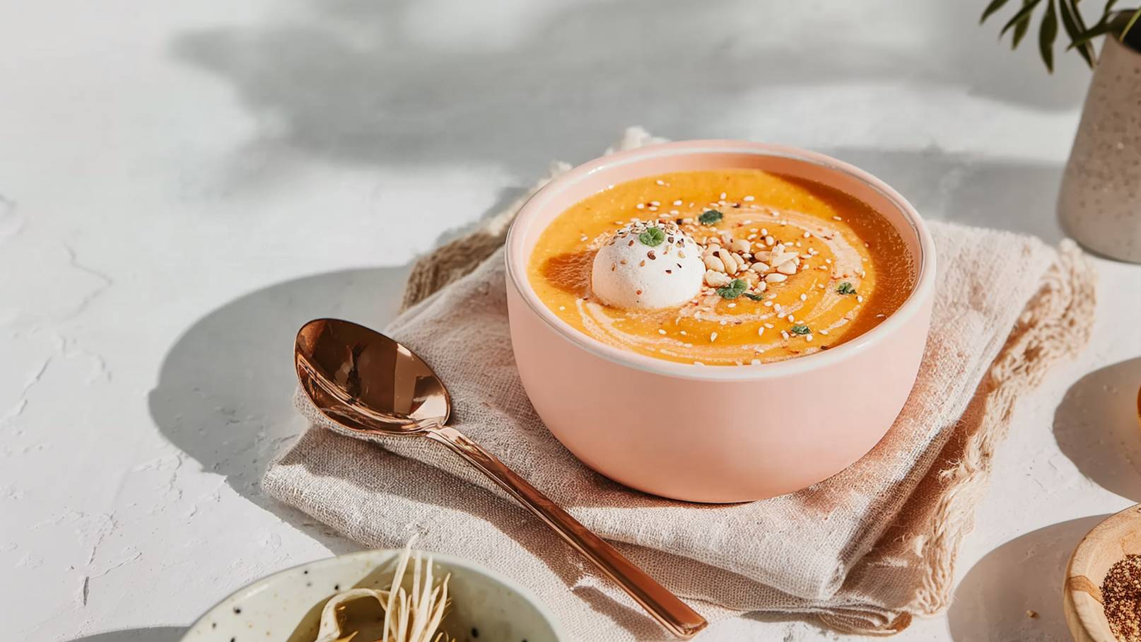 Eine rosa Schüssel mit cremiger Suppe ist auf einem hellen Untergrund platziert. Die Suppe ist mit einer Sahnehaube, Pinienkernen und Kräutern garniert. Neben der Schüssel liegt ein Löffel auf einem Stoffserviette.