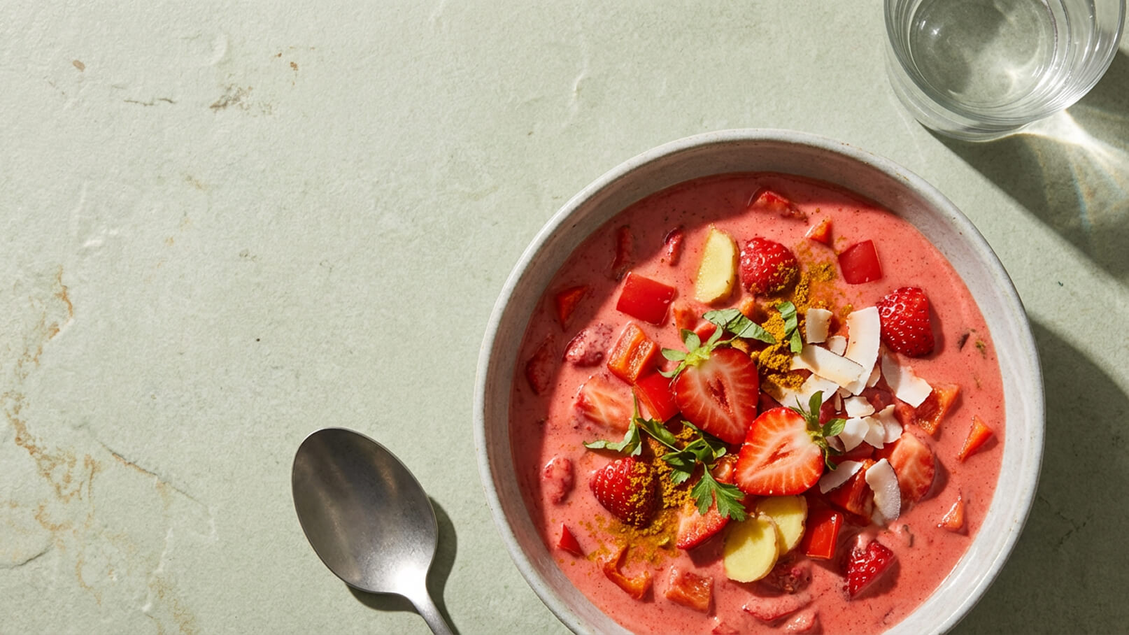 Eine Schüssel mit pinkem Smoothie, garniert mit frischen Erdbeeren, roten Fruchtstücken, Kokosraspeln und grünen Kräutern, steht auf einem hellgrünen Tisch. Daneben liegt ein Löffel und ein Glas Wasser ist im Hintergrund sichtbar.