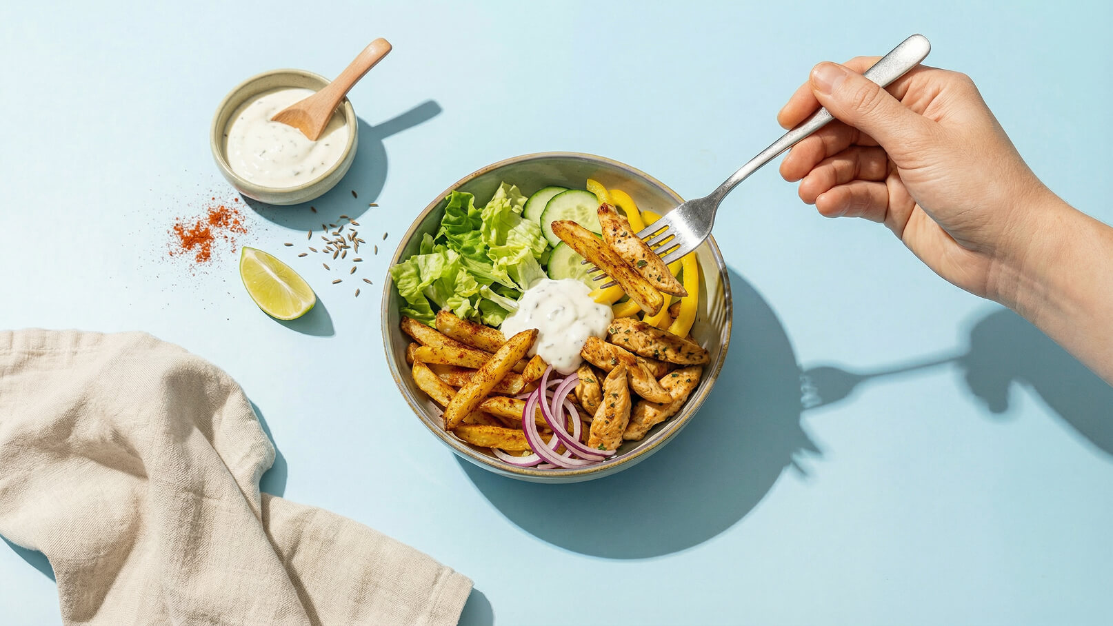 Eine Hand hält eine Gabel, die ein Gericht aus gebratenen Kartoffeln, Hühnchenstreifen, frischen Salatblättern, Gurken, Paprika und einer Joghurtsauce aufnimmt. Neben der Schüssel stehen Limette, Gewürze und eine Schale mit Sauce auf einem hellblauen Hintergrund.