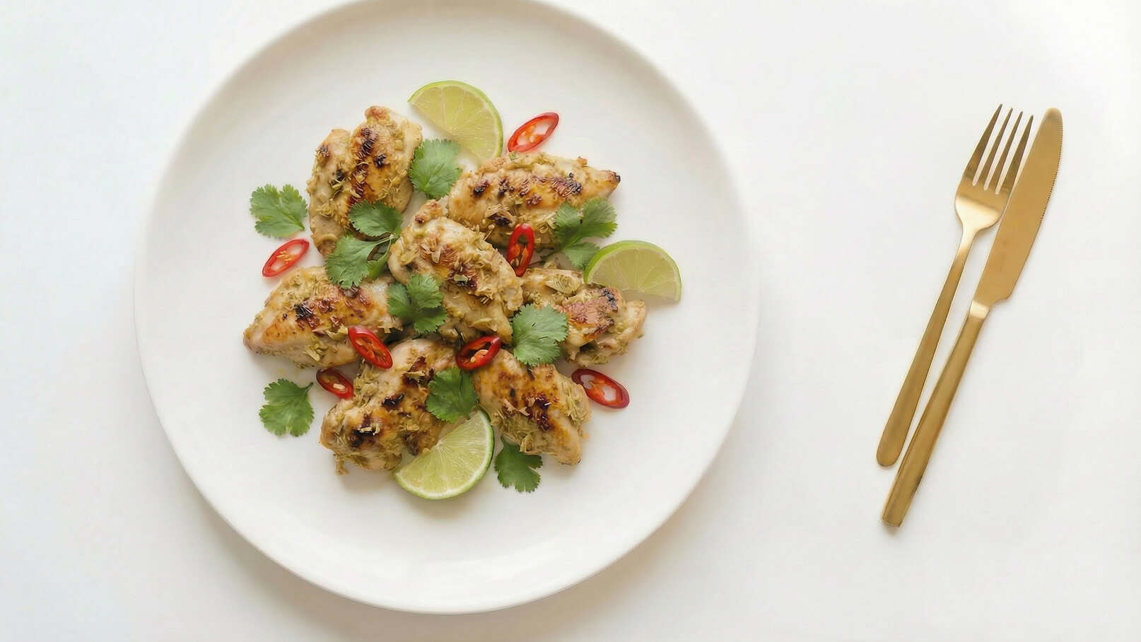 Ein weißer Teller mit gegrillten Hähnchenstücken, garniert mit frischem Koriander, roten Chilischoten und Limettenscheiben. Der Teller liegt auf einer hellen Oberfläche, daneben liegen eine goldene Gabel und ein goldenes Messer.