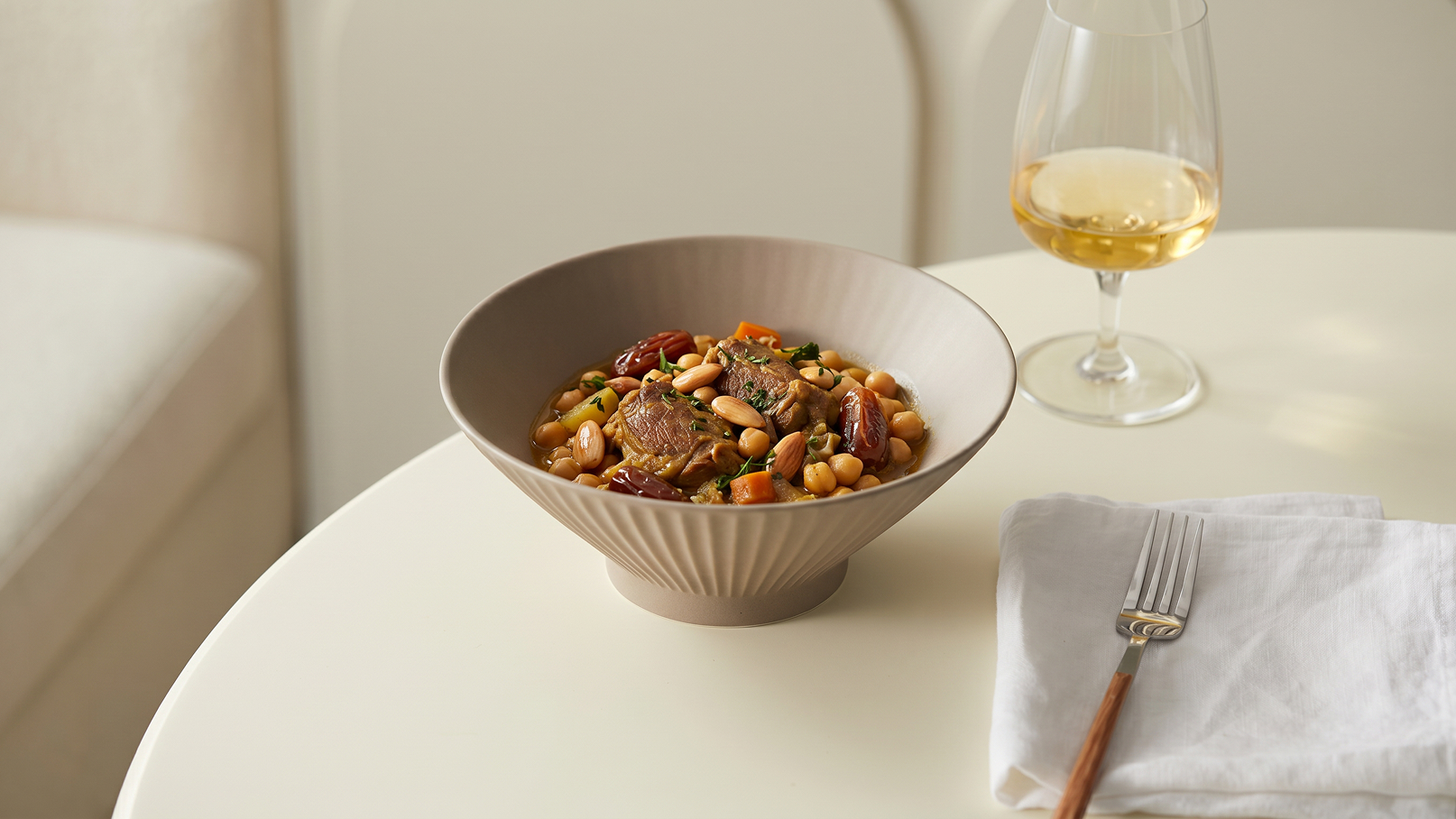 Eine tiefes Schälchen mit einem bunten Gericht, das Fleisch, Gemüse und Bohnen enthält, steht auf einem Tisch. Daneben steht ein Glas mit hellem Getränk und eine Serviette mit Besteck.
