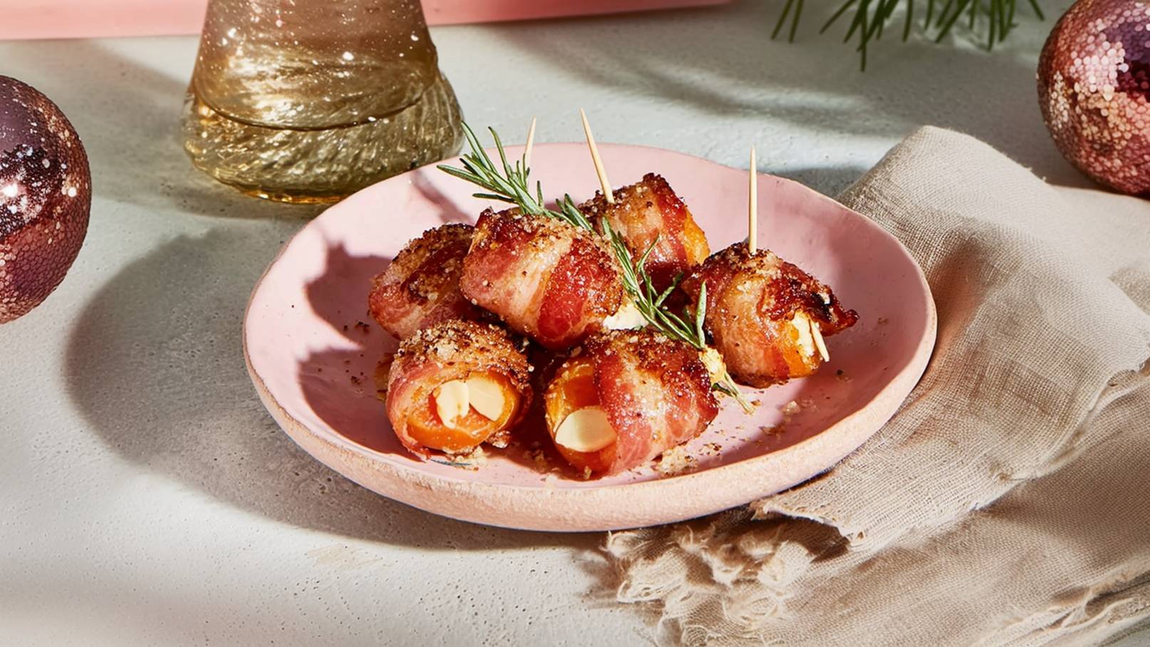 Auf dem Bild ist ein rosa Teller mit mehreren umwickelten Snacks zu sehen, die in Speck gehüllt sind. Die Snacks sind mit einem Zahnstocher gesichert und mit Gewürzen bestreut. Ein Zweig Rosmarin dekoriert die Speisen. Im Hintergrund sind einige weihnachtliche Dekorationselemente sichtbar.