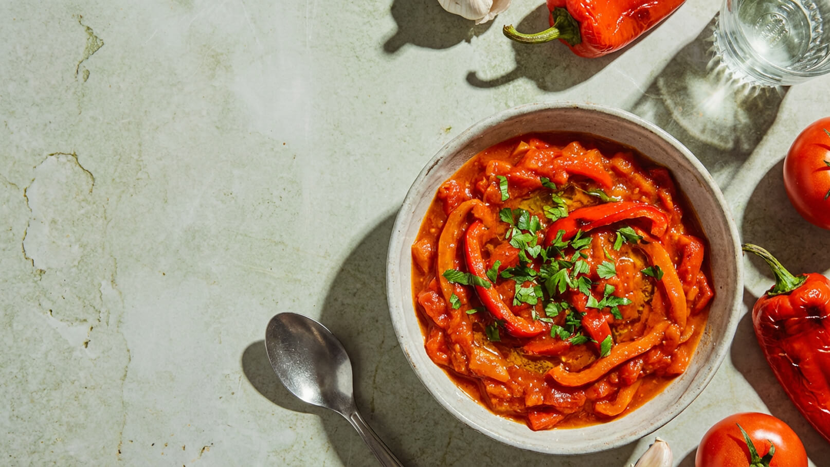 Eine Schüssel mit rotem Paprika-Eintopf, garniert mit frischer Petersilie. Auf dem Tisch liegen einige frische Tomaten, Paprikaschoten und eine Knoblauchzehe. Ein Löffel ist neben der Schüssel positioniert.