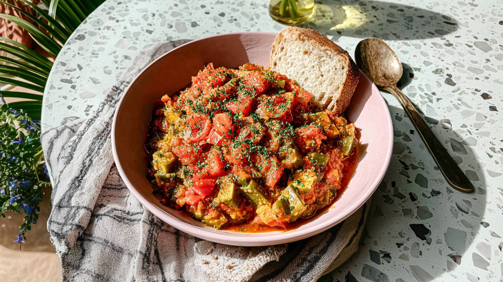 Eine pinke Schüssel mit einem bunten Gemüsegericht, hauptsächlich aus Tomaten und Paprika, ist auf einem Tisch platziert. Neben der Schüssel liegt ein Stück Brot. Im Hintergrund sind ein Löffel und eine Serviette sichtbar.