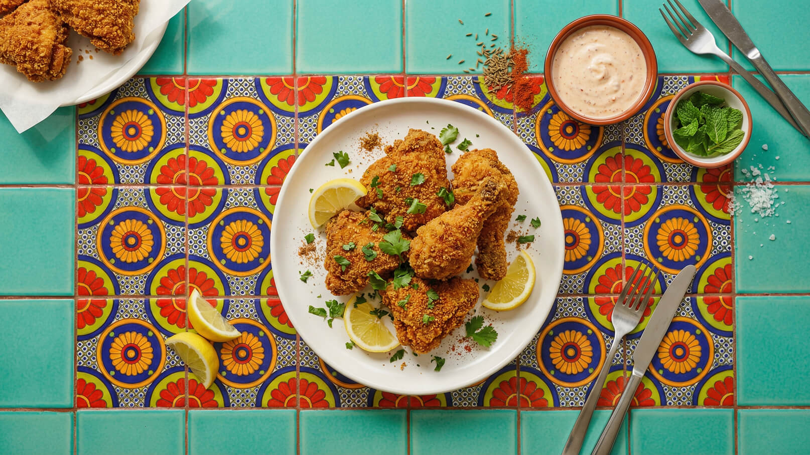Ein Teller mit knusprig gebratenem Hähnchen, garniert mit frischen Kräutern und Zitronenscheiben. Der Hintergrund zeigt eine bunt gemusterte Fliese. Neben dem Teller steht eine kleine Schüssel mit einer cremigen Sauce und frischen Minzblättern.