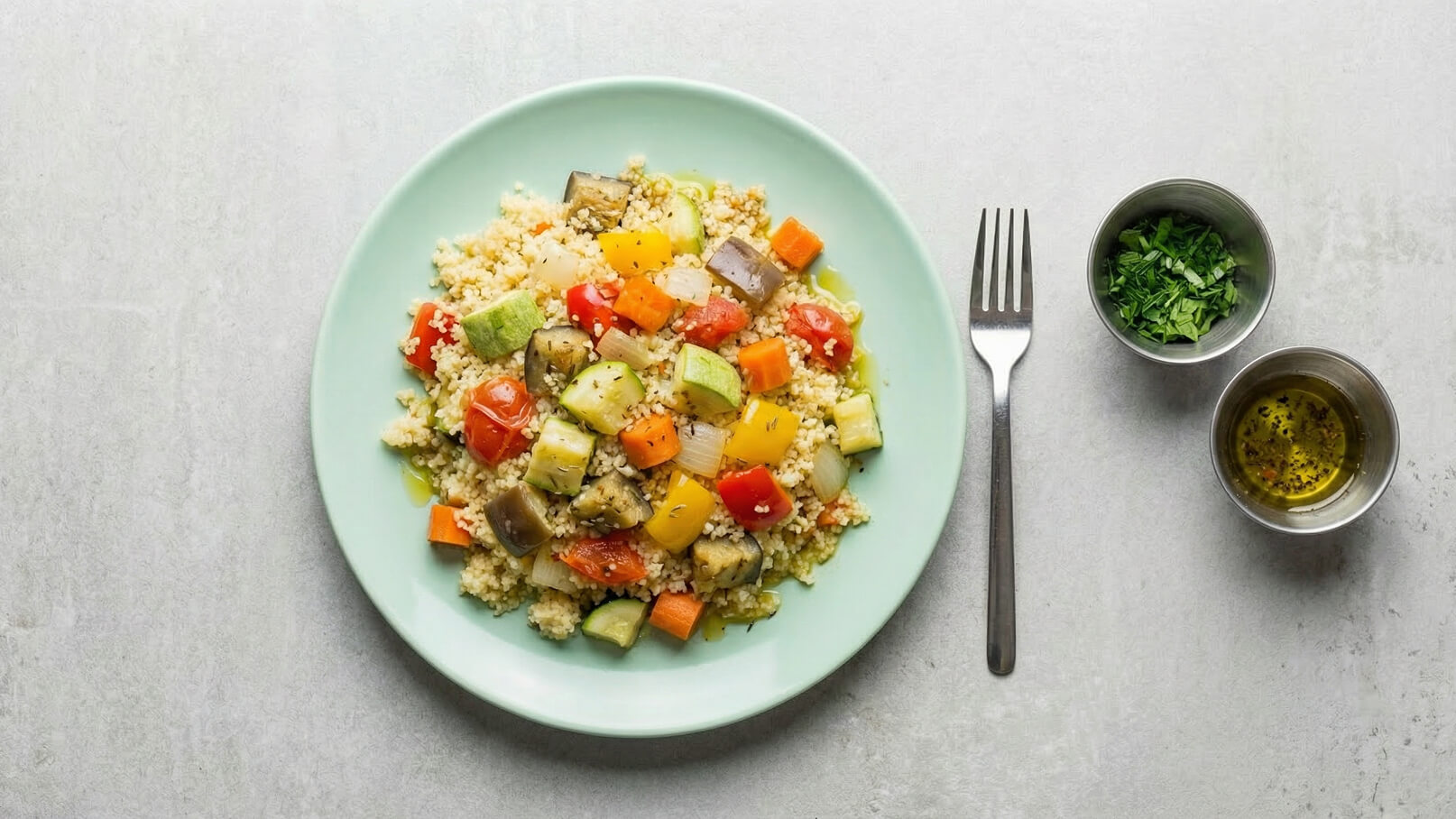 Ein Teller mit Couscous, garniert mit buntem Gemüse wie Tomaten, Paprika, Zucchini und Karotten. Daneben befinden sich zwei Schalen mit frischen Kräutern und einer Ölmischung. Ein Besteckstück liegt auf der rechten Seite des Tellers.