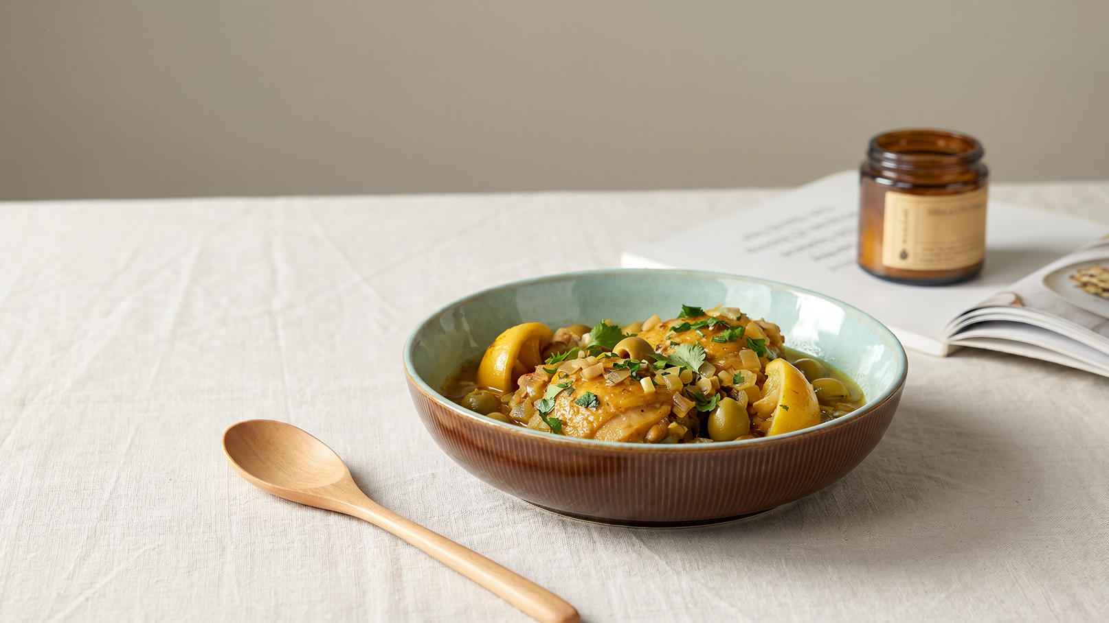 Eine braune Schüssel enthält ein Gericht mit Hühnchen, Äpfeln und Kräutern, serviert auf einem hellen Tisch. Neben der Schüssel liegt ein Holzlöffel und im Hintergrund ist ein Buch sowie ein Glas sichtbar.