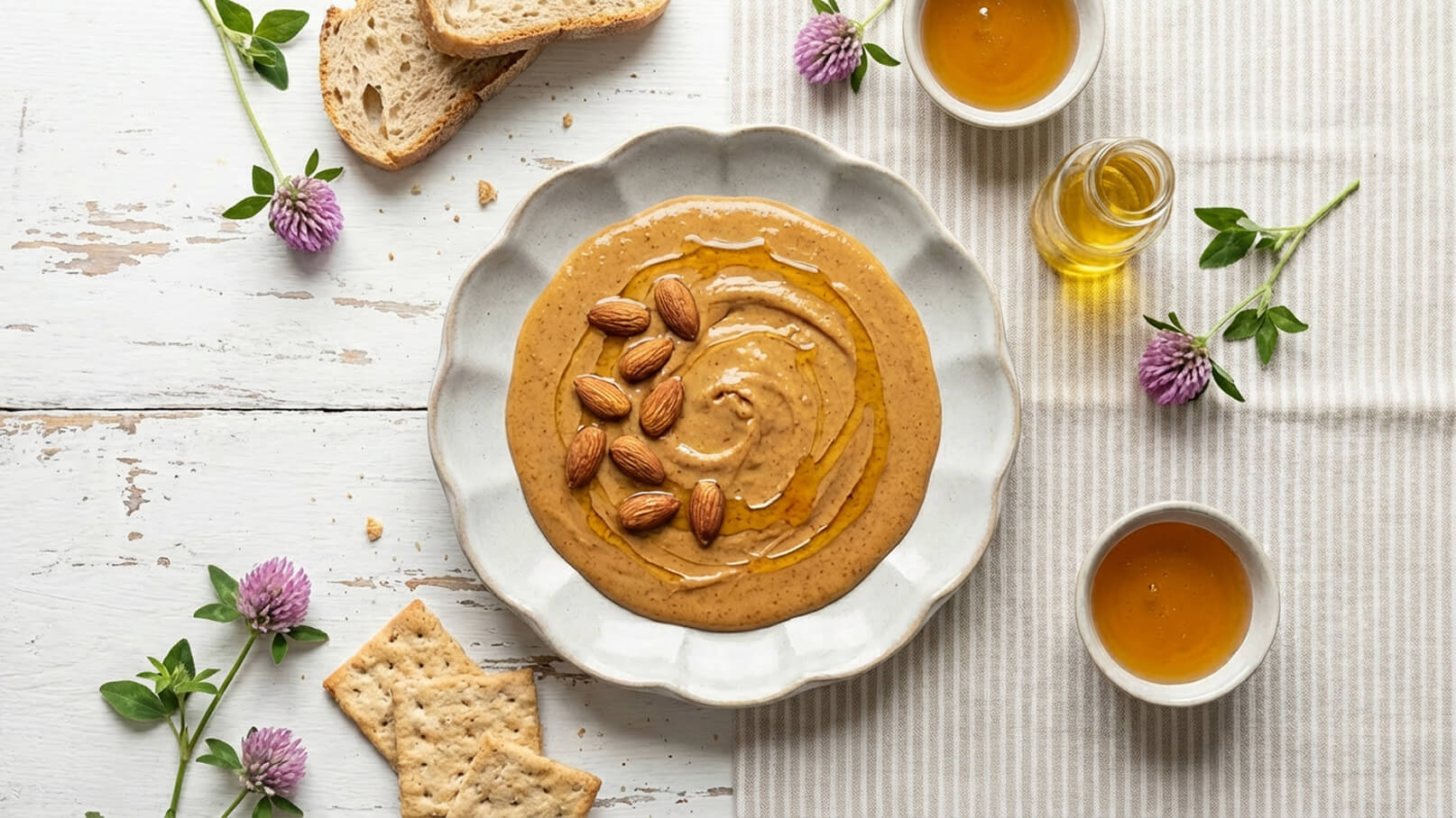 Eine flache, runde Schüssel mit cremiger Erdnussbutter, garniert mit Mandeln und einem Schuss Honig. Daneben liegen einige Stücke Brot und ausgebrochene Crackern. Die Unterlage ist gestreift, mit frischen Kräutern und einer lila Blüte dekoriert.