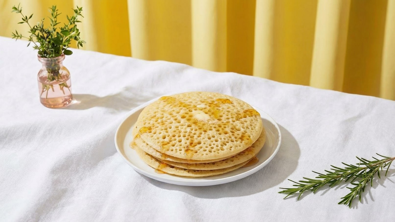 Ein Teller mit drei Stapeln Crumpets, die leicht goldbraun sind und etwas Butter und Honig darauf haben. Der Teller steht auf einem weißen Tuch. Im Hintergrund ist ein gelbes Vorhang und eine kleine Glasvase mit frischen Kräutern zu sehen.