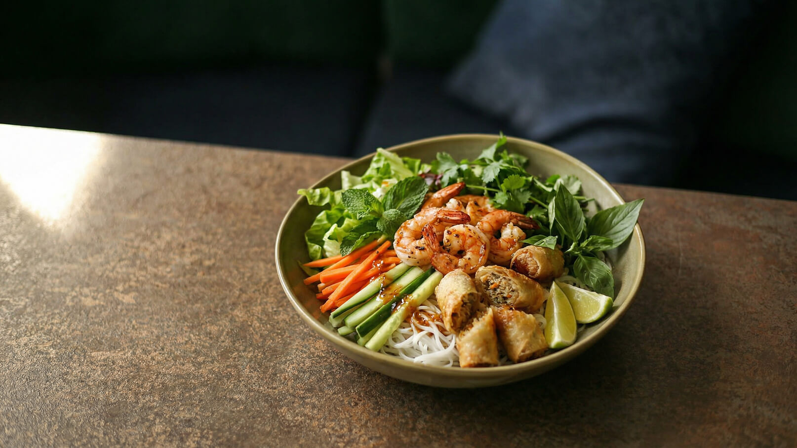 Eine Schüssel mit einem bunten Salat, der Garnelen, Reisnudeln, frischen Kräutern, Karottenstreifen, Gurkenscheiben und gebratenen Frühlingsrollen enthält. Ein Stück Limette ist ebenfalls enthalten. Der Salat ist ansprechend angerichtet und zeigt eine Vielfalt an Farben und Texturen.