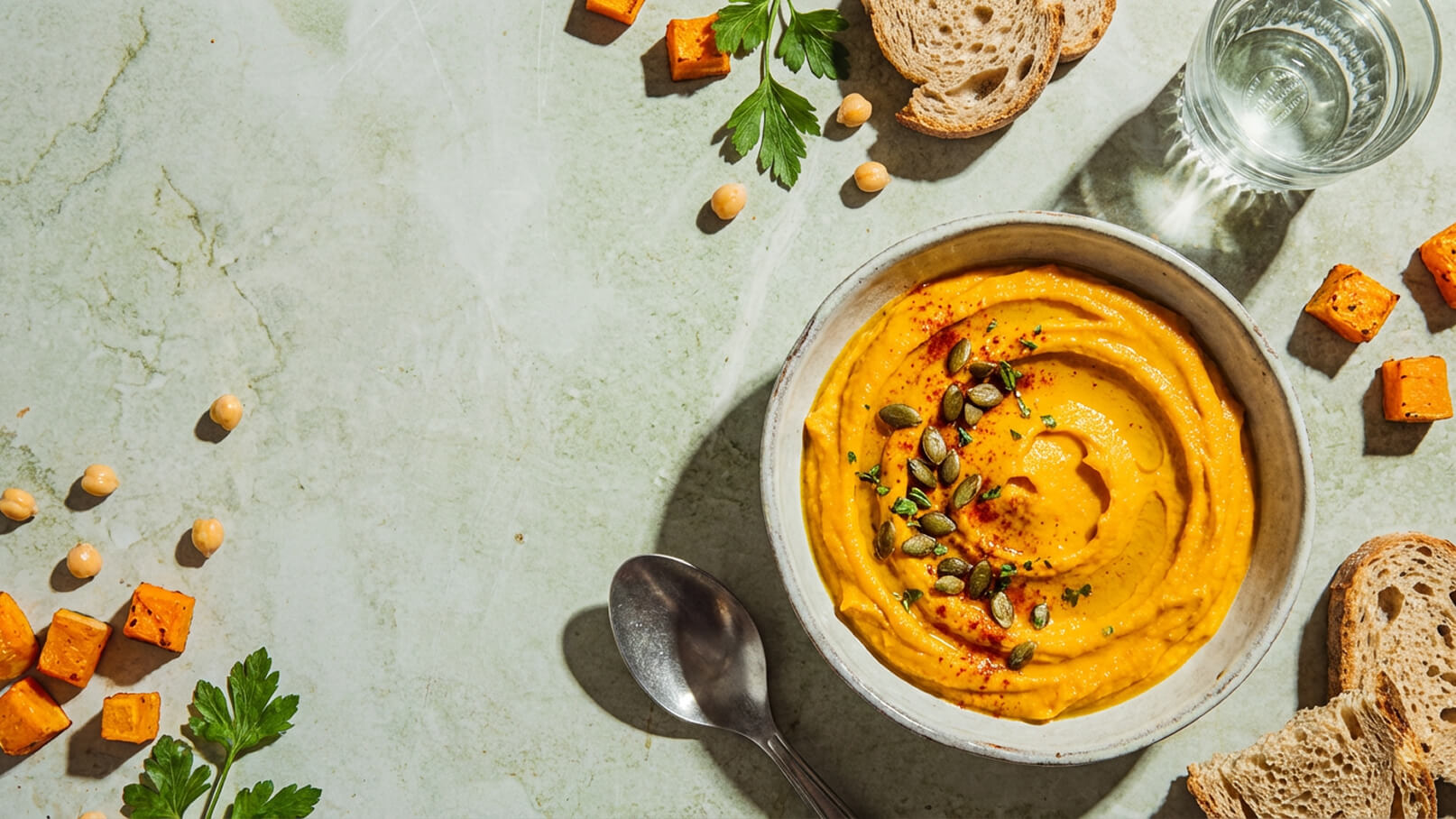 Eine Schüssel mit cremiger, orangefarbener Suppe, garniert mit Kürbiskernen und einem Hauch Paprika. Daneben liegen frisches Brot, einige Stücke geschnittenes Gemüse und ein Glas Wasser auf einem marmorierten Tisch.