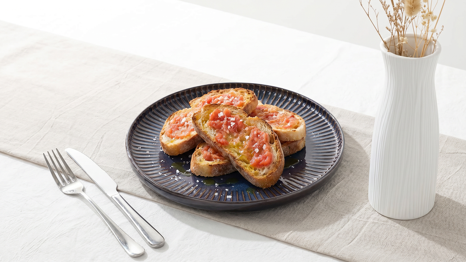Ein dunkler Teller mit mehreren Scheiben geröstetem Brot, belegt mit gewürfelten Tomaten und Kräutern. Neben dem Teller liegen Besteck und ein blühender Vase. Der Tisch ist hell und einfach gedeckt.