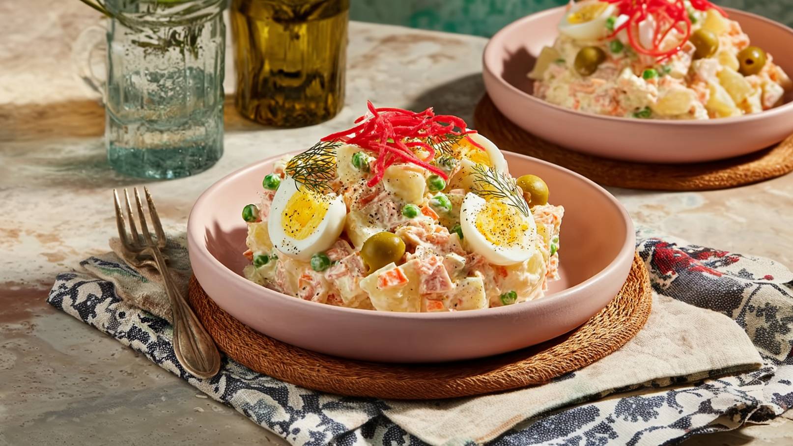 Zwei Teller mit Kartoffelsalat sind auf einem Tisch angerichtet. Der Salat enthält Kartoffeln, Erbsen, Gewürzgurken und Eier, garniert mit roter Zwiebel und Dill. Das Geschirr ist rosa, umgeben von einer geflochtenen Unterlage und einem dekorativen Stoff.