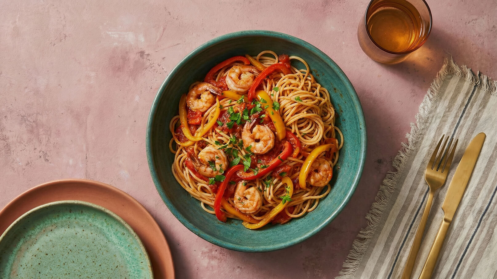 Ein tiefes, türkisfarbenes Schüssel enthält Spaghetti mit Garnelen, roten und gelben Paprikastreifen sowie frischer Petersilie. Daneben liegen ein goldenes Besteck und eine gestreifte Serviette auf einem rosa Untergrund.