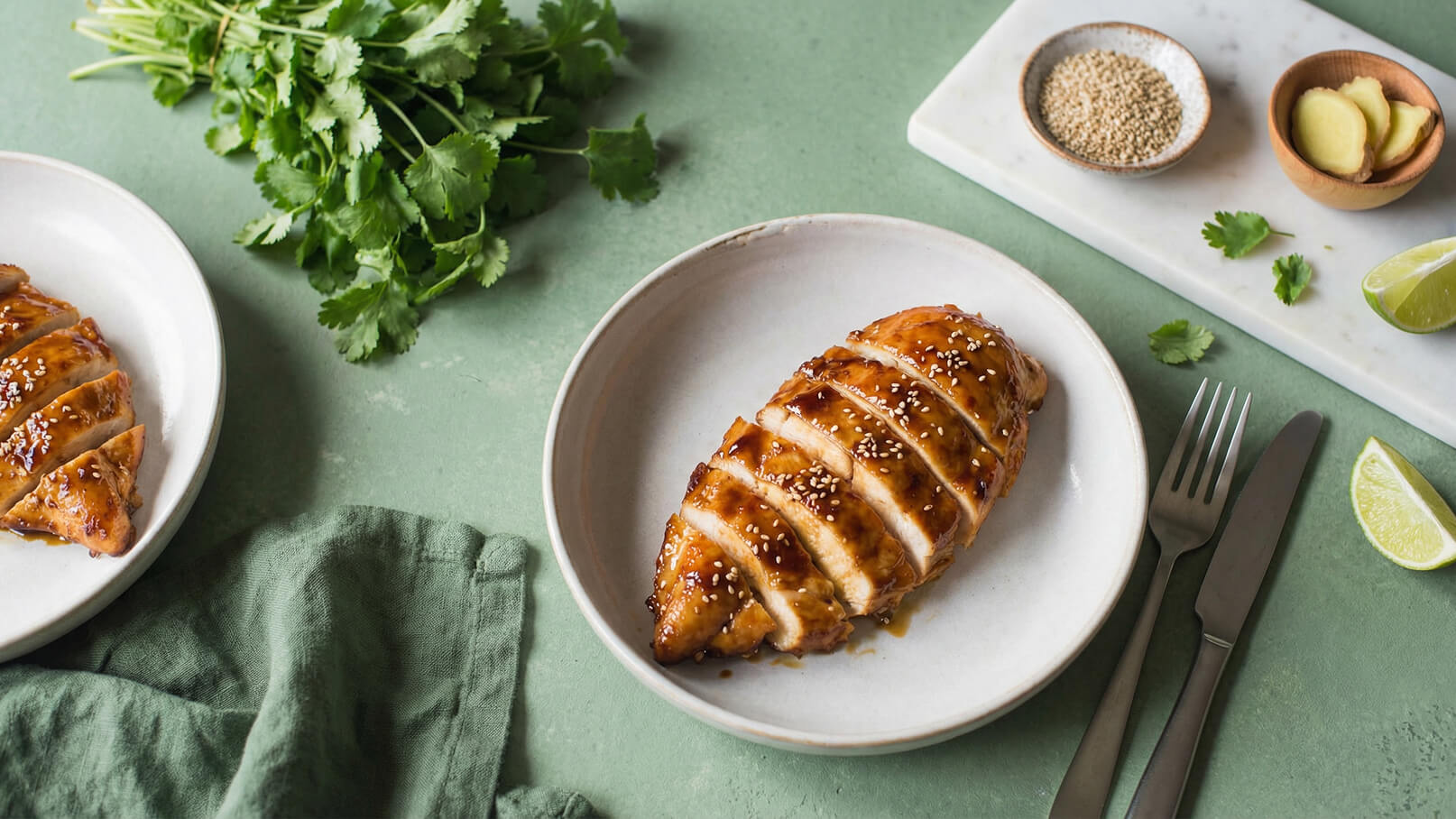 Ein Teller mit geschnittenem, glasierter Hühnchenbrust, bestreut mit Sesamsamen. Daneben frische Korianderblätter und ein kleines Brett mit einer Schüssel Sesam und Ingwer sowie einer Limette. Der Hintergrund ist in sanften Grüntönen gehalten.