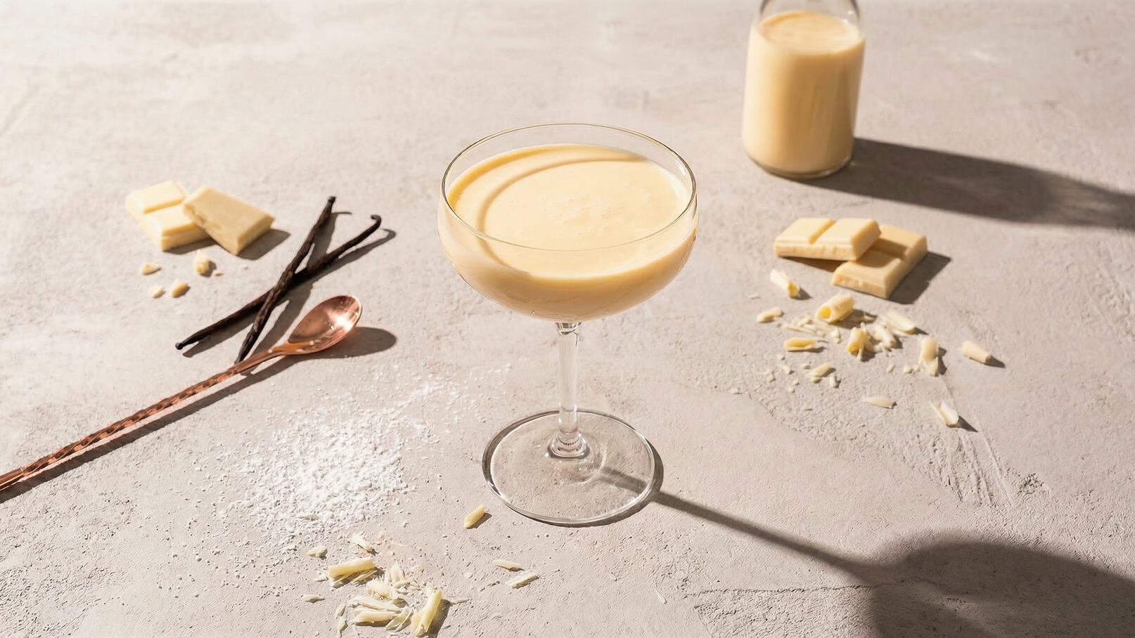 Ein elegantes Glas mit einer cremigen Vanille-Speise steht auf einer grauen Oberfläche. Um das Glas sind weiße Schokolade, Vanilleschoten und eine kupferne Löffel dekorativ angeordnet. Im Hintergrund ist eine Glasflasche mit einer ähnlichen Vanille-Creme zu sehen.