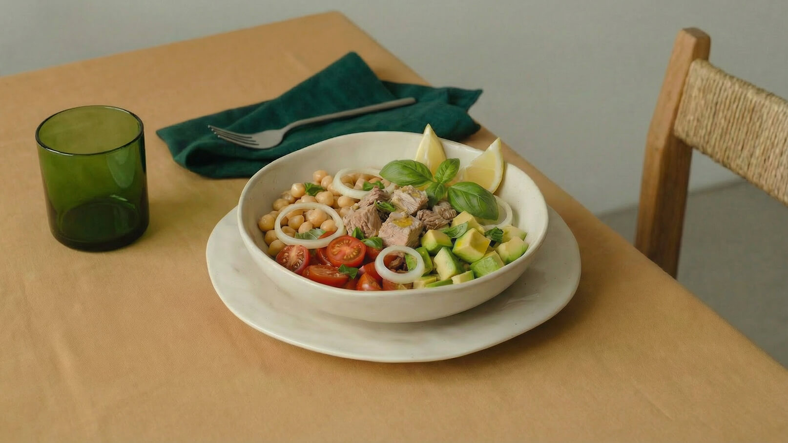 Eine helle Schüssel mit einem bunten Salat aus Kichererbsen, Tomatenstücken, Avocado und Tunfisch, garniert mit frischem Basilikum und Zitronenscheiben. Daneben steht ein grünes Glas und eine Serviette auf einem hellen Tisch.