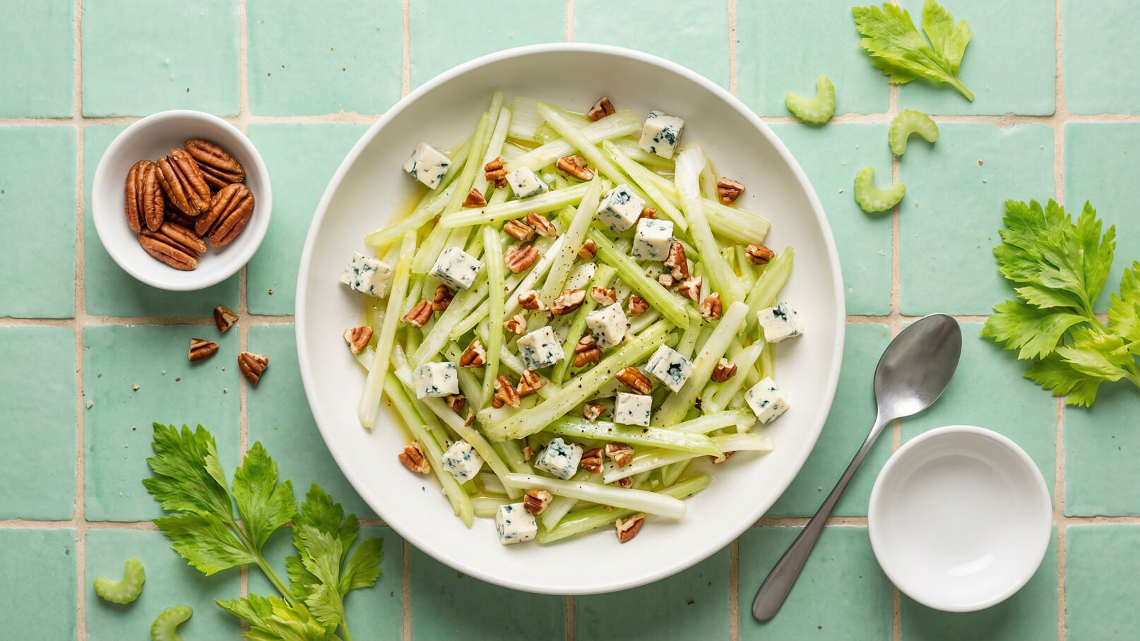 Ein runder, weißer Teller enthält einen Salat aus zarten Selleriestangen, gewürfeltem Blauschimmelkäse und gehackten Pekannüssen. Der Teller steht auf einem hellgrünen Fliesenuntergrund. Neben dem Teller befinden sich eine Schüssel mit Pekannüssen und ein Löffel.