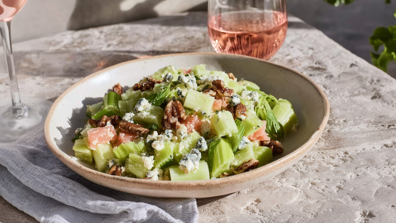 Ein Teller mit frischem Salat, bestehend aus knusprigem Sellerie, kleinen Stücken Lachs, gewürfeltem Blauschimmelkäse und gerösteten Pekannüssen. Der Salat wird auf einem Tisch serviert, neben einem Glas rosé Farbgetränk.