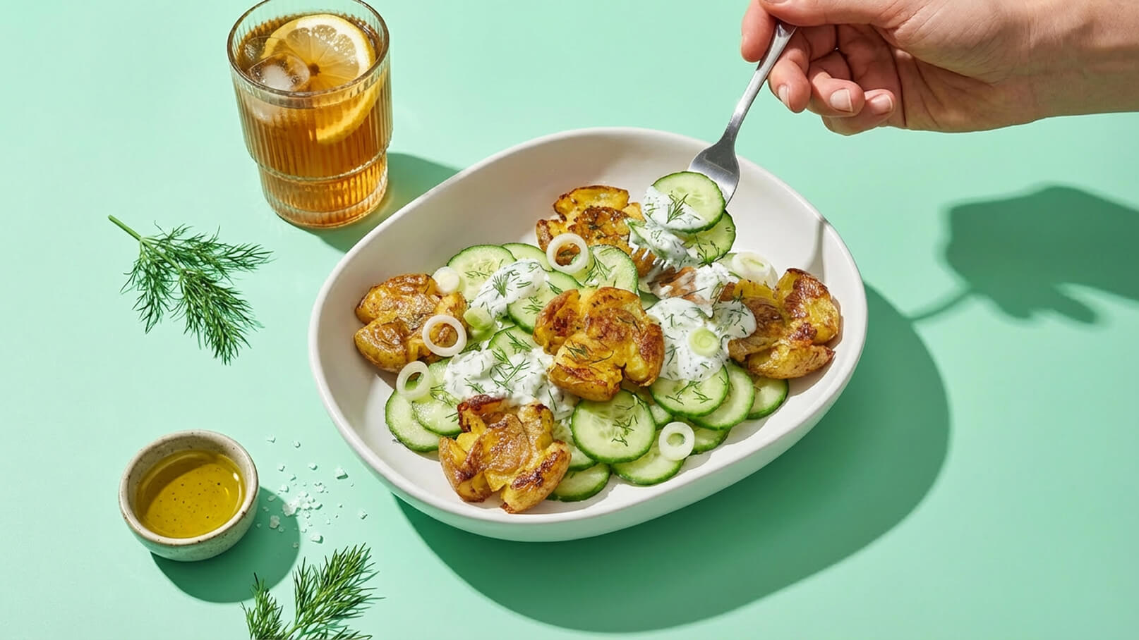 Eine Hand hält eine Gabel und nimmt einen Bissen von einem Teller mit Gurkenscheiben, knusprigen Kartoffeln und einer Kräuterjoghurtsoße. Neben dem Teller steht ein Glas mit einem Getränk und einer Zitronenscheibe. Der Hintergrund ist mintgrün.