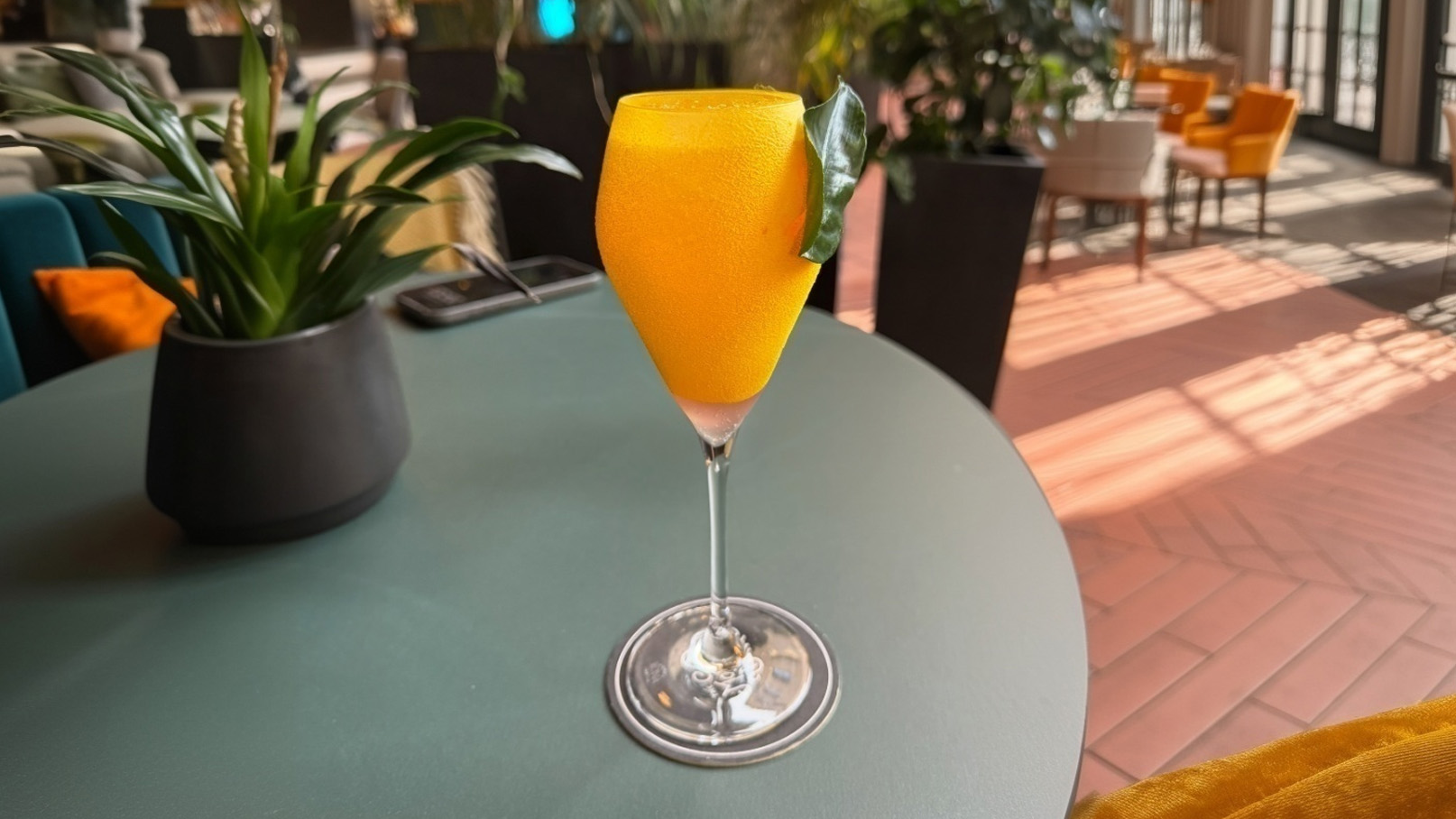 Ein Glas mit einem gelben Cocktail steht auf einem grünen Tisch. Der Drink hat eine frostige Oberfläche und ist mit einem Blatt dekoriert. Im Hintergrund sind verschwommene Möbel und Pflanzen sichtbar, die eine gemütliche Atmosphäre schaffen.